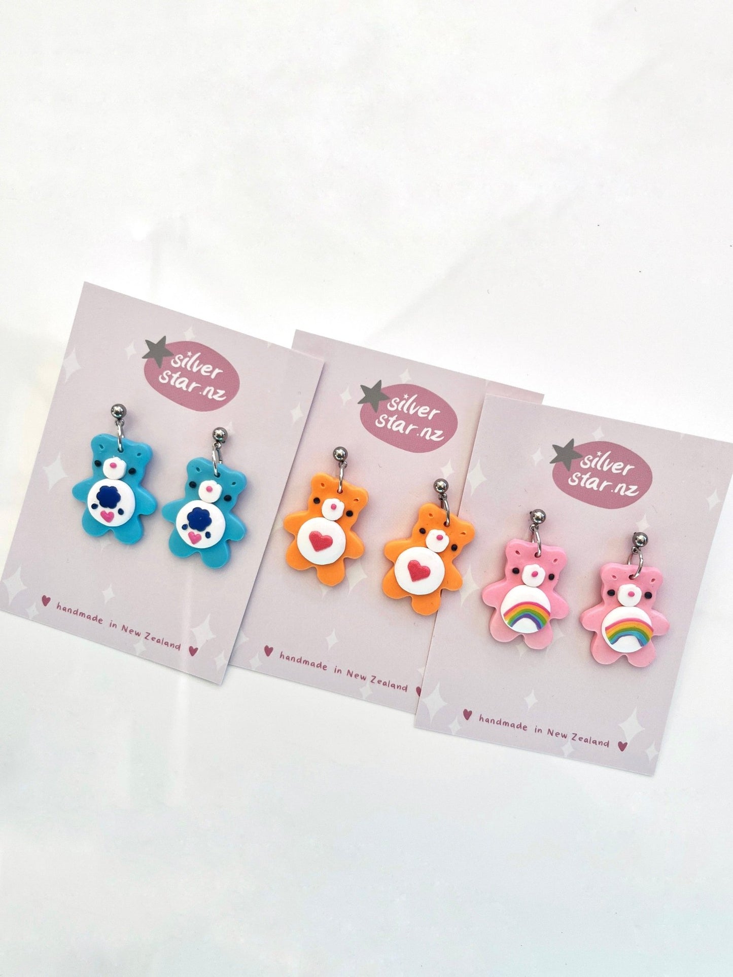 Four pairs of Care Bear Polymer Clay Earrings are showcased on cards labeled with the brand silverstar.nz. The bear-shaped earrings feature designs in blue, orange, and pink, with options for hearts or rainbows on their bellies. These distinctive pieces are lovingly handmade in New Zealand.