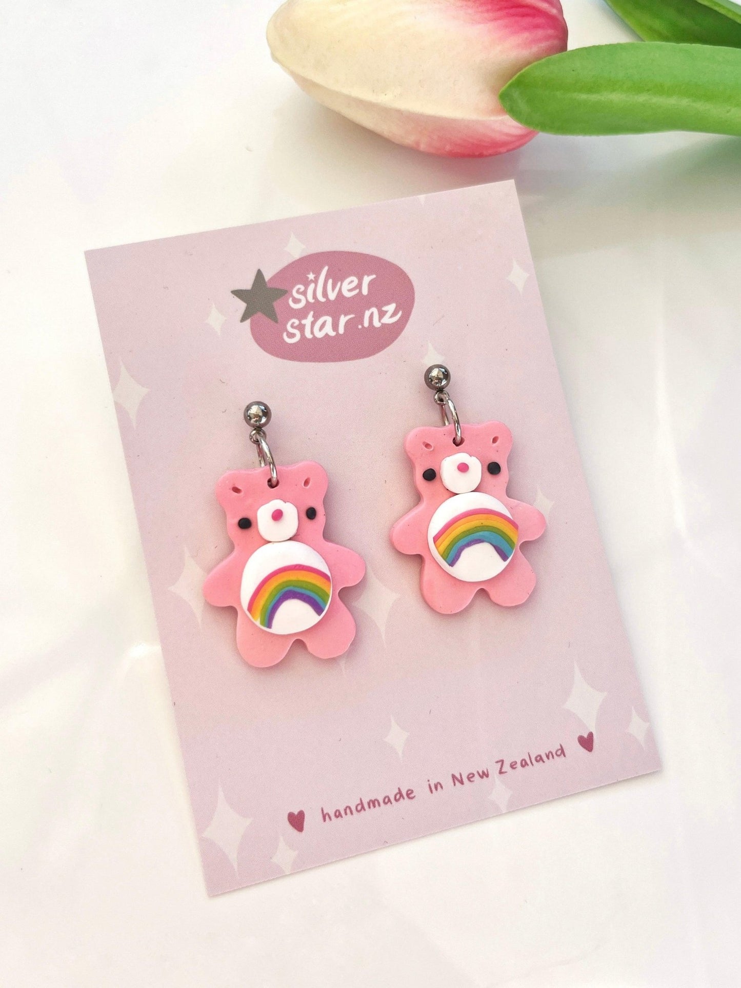 The Care Bear Polymer Clay Earrings, featuring pink bear shapes with rainbow designs, are showcased on a card branded with silverstar.nz. A nearby pink tulip complements the display, while the card emphasizes their handmade and unique design, crafted in New Zealand.