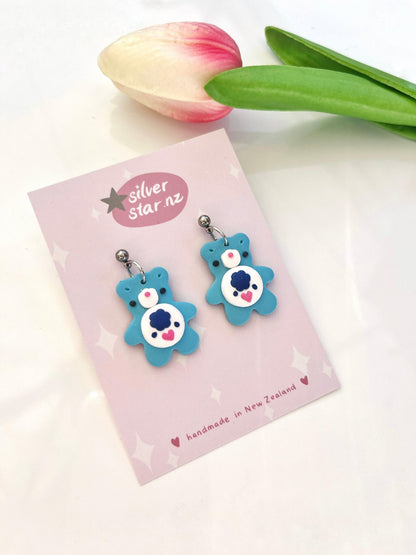 Displayed on an elegantly branded card from silverstar.nz, the Care Bear Polymer Clay Earrings, featuring intricate heart and face details, showcase a delightful blue bear design. A note highlights their handmade craftsmanship in New Zealand. To enhance the display's charm, a pink and white tulip is placed nearby.