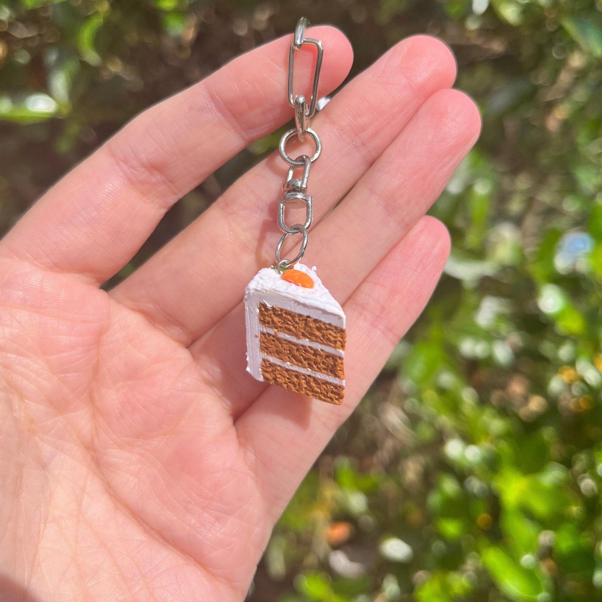 A hand showcases the Carrot Cake Polymer Clay Keyring by silverstar.nz, a distinctive accessory featuring a miniature slice of cake charm. Made from handmade polymer clay, it mimics a layered cake with white frosting and an orange accent, all set against a softly blurred green backdrop.