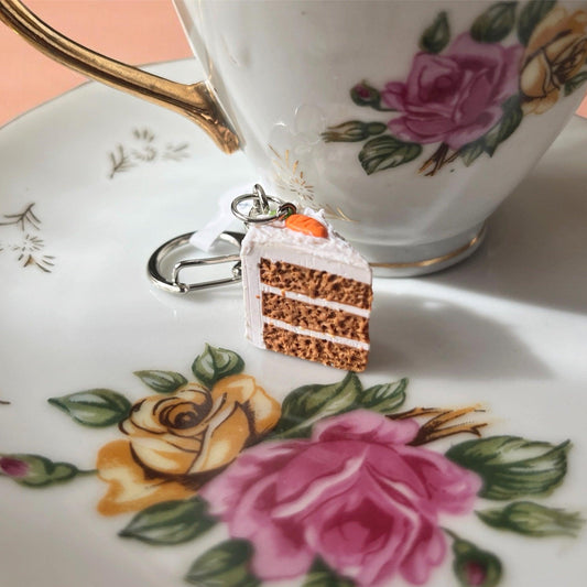 The Carrot Cake Polymer Clay Keyring by silverstar.nz rests on a floral-patterned plate beside a porcelain teacup adorned with gold accents. This handmade charm showcases layers of brown cake and white frosting, topped with a miniature carrot, and features a distinctive funky design paired with a durable keyring clasp.