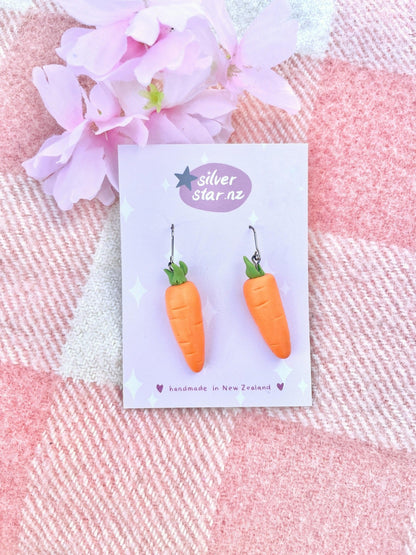 Carrot-shaped polymer clay earrings from silverstar.nz on a display card, featuring a pink and white plaid fabric background. The card is adorned with pink flowers in the top left corner. These handmade earrings, crafted in New Zealand, come with stainless steel hooks.