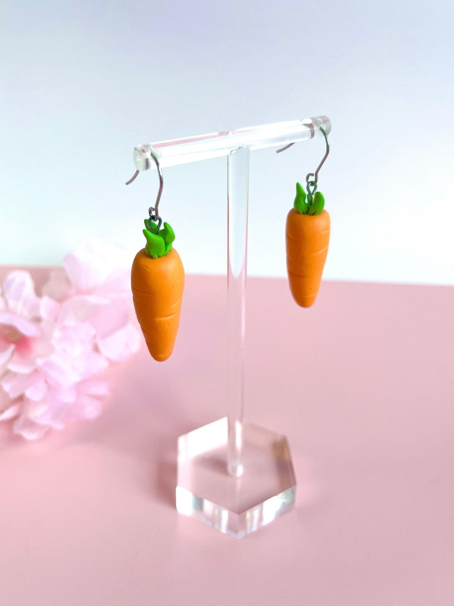 Introducing the Carrot Polymer Clay Earrings by silverstar.nz, a delightful pair featuring charming carrot shapes with green tops. These earrings are meticulously crafted from polymer clay and fitted with stainless steel hooks. They are elegantly displayed on a transparent stand against a soft pink surface scattered with light pink flowers, showcasing their exquisite handmade appeal.