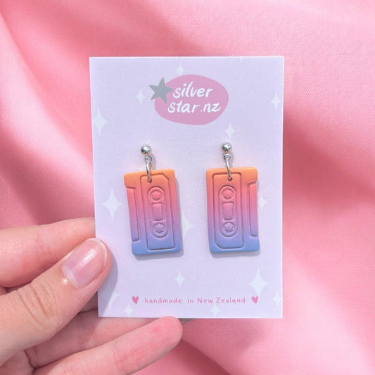 A hand holds a card showcasing the Casette Polymer Clay Earrings by silverstar.nz, uniquely designed as funky retro cassette tapes in an orange and purple gradient. These earrings are handmade in New Zealand, featuring stainless steel hooks and displayed against a soft pink fabric background.