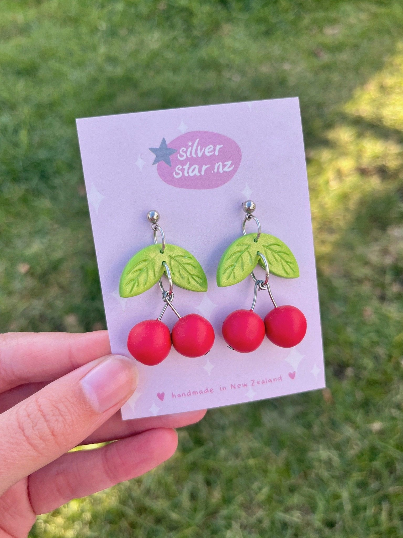 A hand is holding a card displaying the Cherry Polymer Clay Earrings from silverstar.nz, showcasing their handmade origin in New Zealand. These distinctively designed earrings feature red cherries and green leaves against a light pink background, set amidst a backdrop of vibrant green grass.