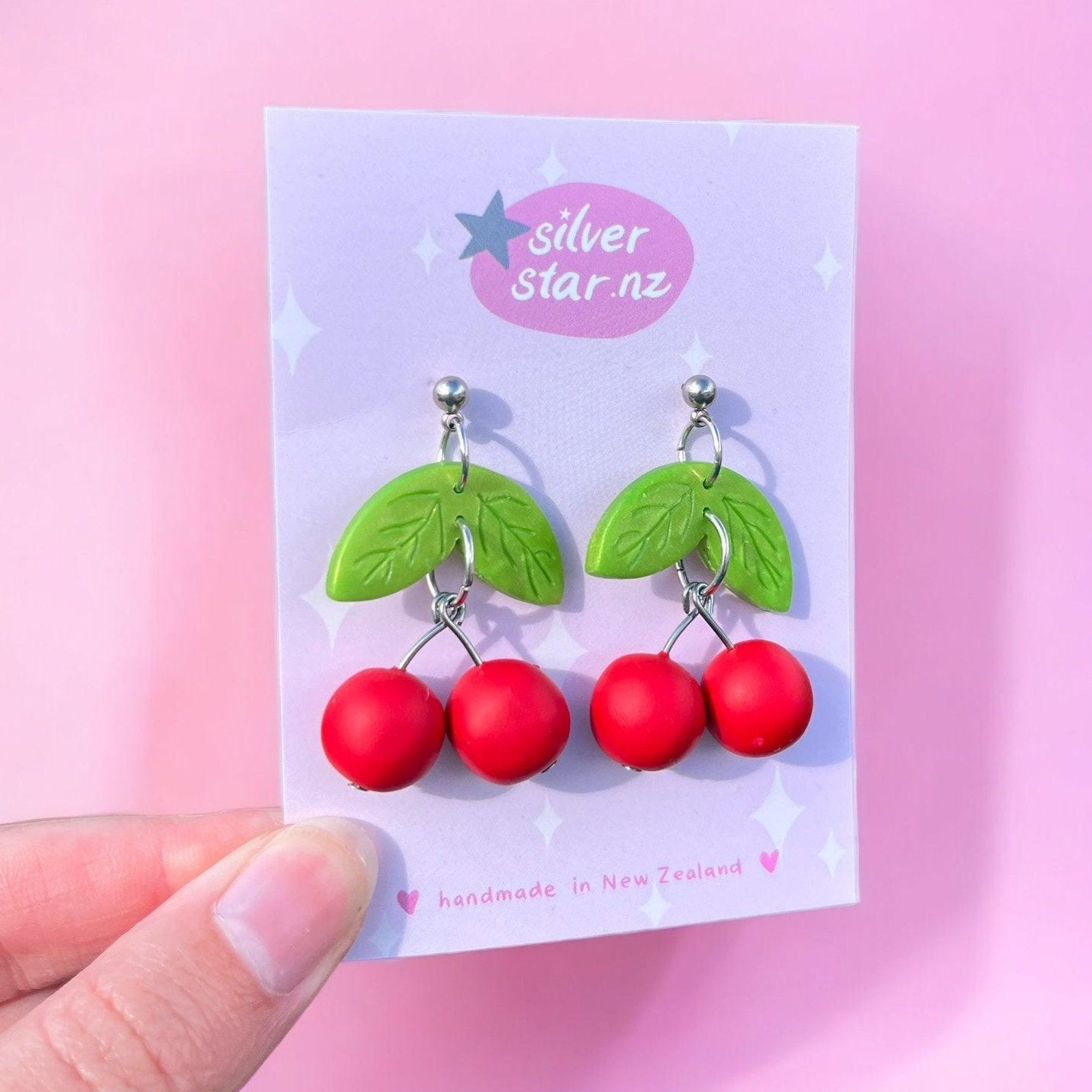 The Cherry Polymer Clay Earrings by silverstar.nz are a distinctive handmade design featuring red cherries with green leaves. These earrings are crafted from polymer clay and elegantly hang on a card displaying the silverstar.nz label, set against a pink backdrop, and gracefully showcased by a hand positioned in the lower left corner.