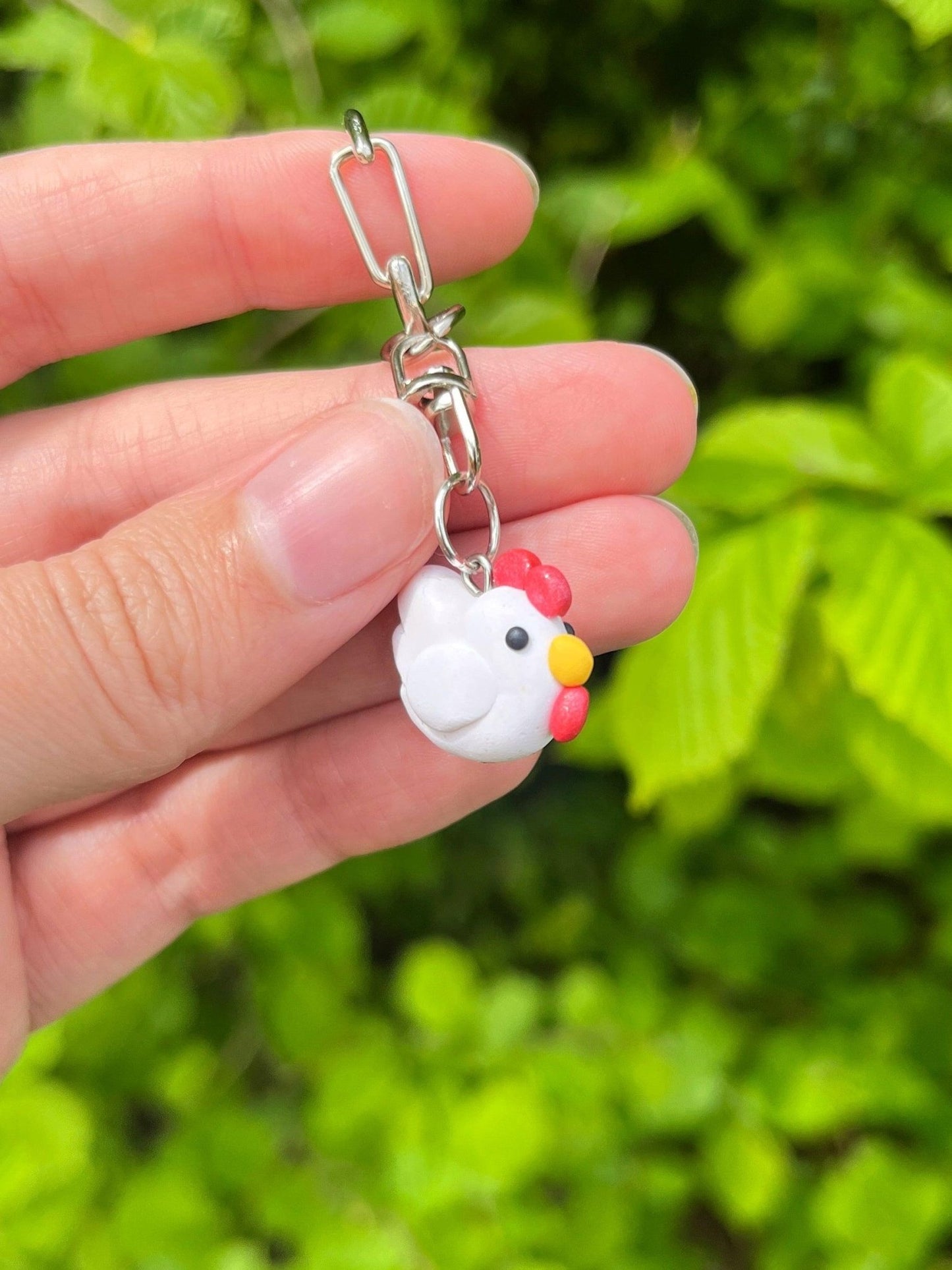 A hand holds the Chicken Polymer Clay Keyring by silverstar.nz, showcasing a small and unique design with a cute white chicken, accented by a red comb and yellow beak, and completed with a sleek silver keyring clasp against a backdrop of green leaves.