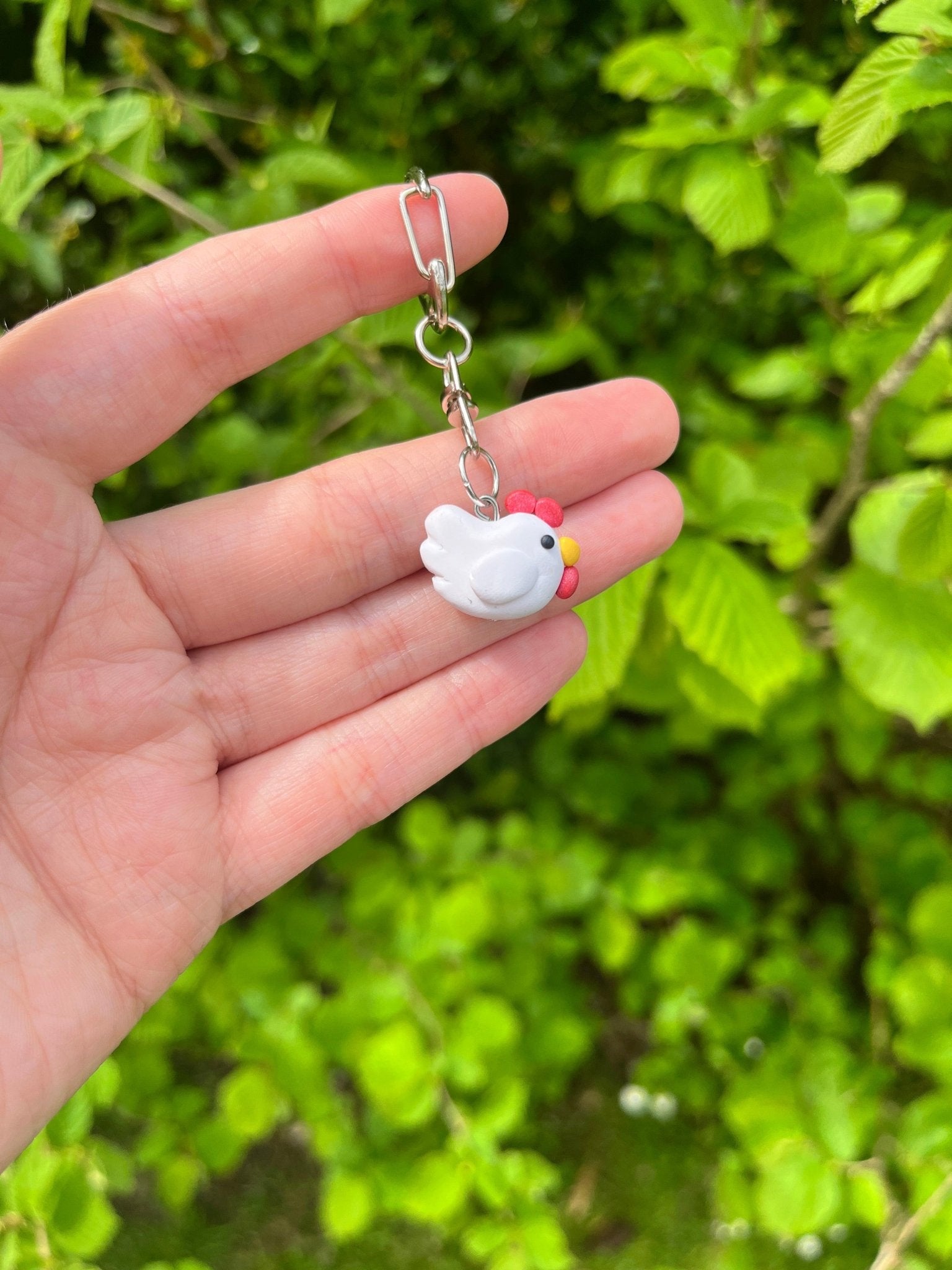 A hand showcases the Chicken Polymer Clay Keyring from silverstar.nz, highlighting a distinctive small white bird with pink accents. This keychain, equipped with a silver keyring clasp, is beautifully presented against a lush green leafy bush backdrop.