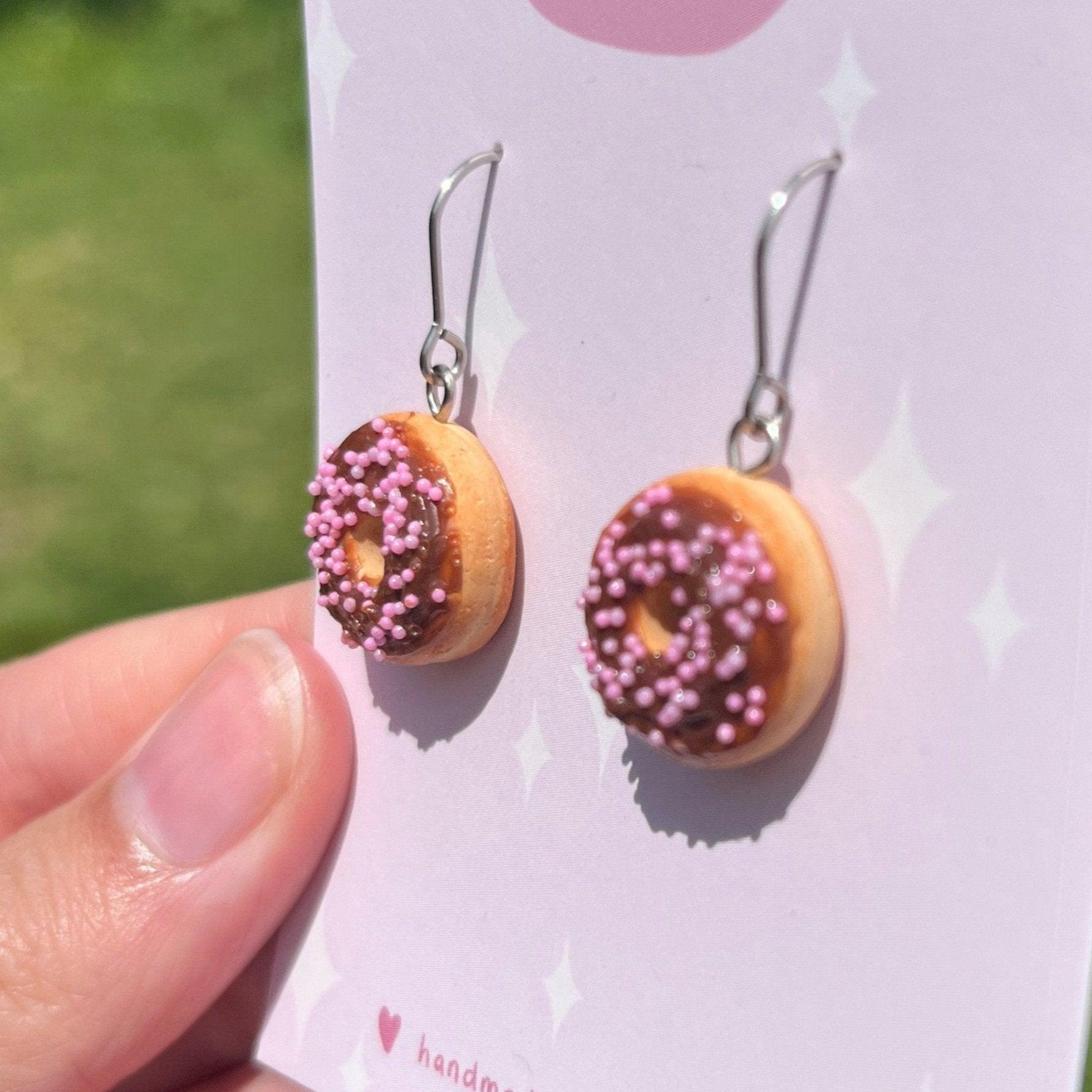 A close-up shot of the Choc Iced Donut Polymer Clay Earrings by silverstar.nz shows them held against a green background. These handmade earrings feature chocolate icing with pink sprinkles and are attached to a subtly star-patterned card. They include durable stainless steel hooks.