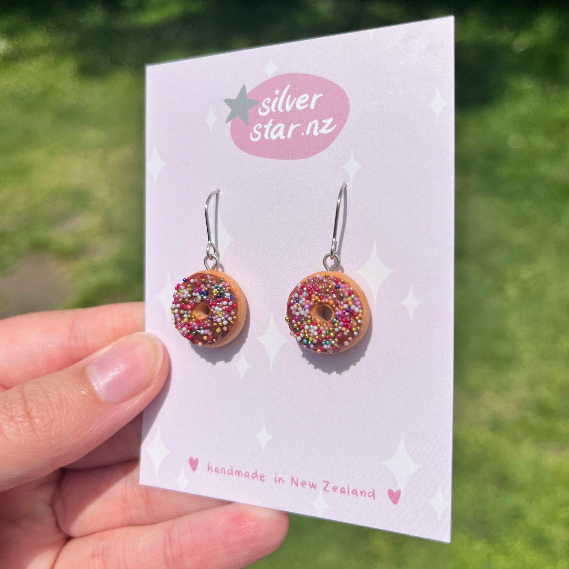 A close-up shows a hand holding the Choc Iced Donut Polymer Clay Earrings, designed with colorful sprinkles and mounted on a card labeled silverstar.nz. These earrings feature stainless steel hooks. The background is a blurred outdoor scene with greenery.