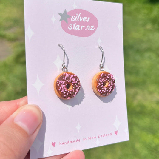 Displayed on a card, the Choc Iced Donut Polymer Clay Earrings are crafted to resemble mini chocolate donuts with pink icing and sprinkles. The card, labeled with the brand name silverstar.nz, indicates they are handmade in New Zealand and feature stainless steel hooks. In the background, a grassy area is illuminated by sunlight.