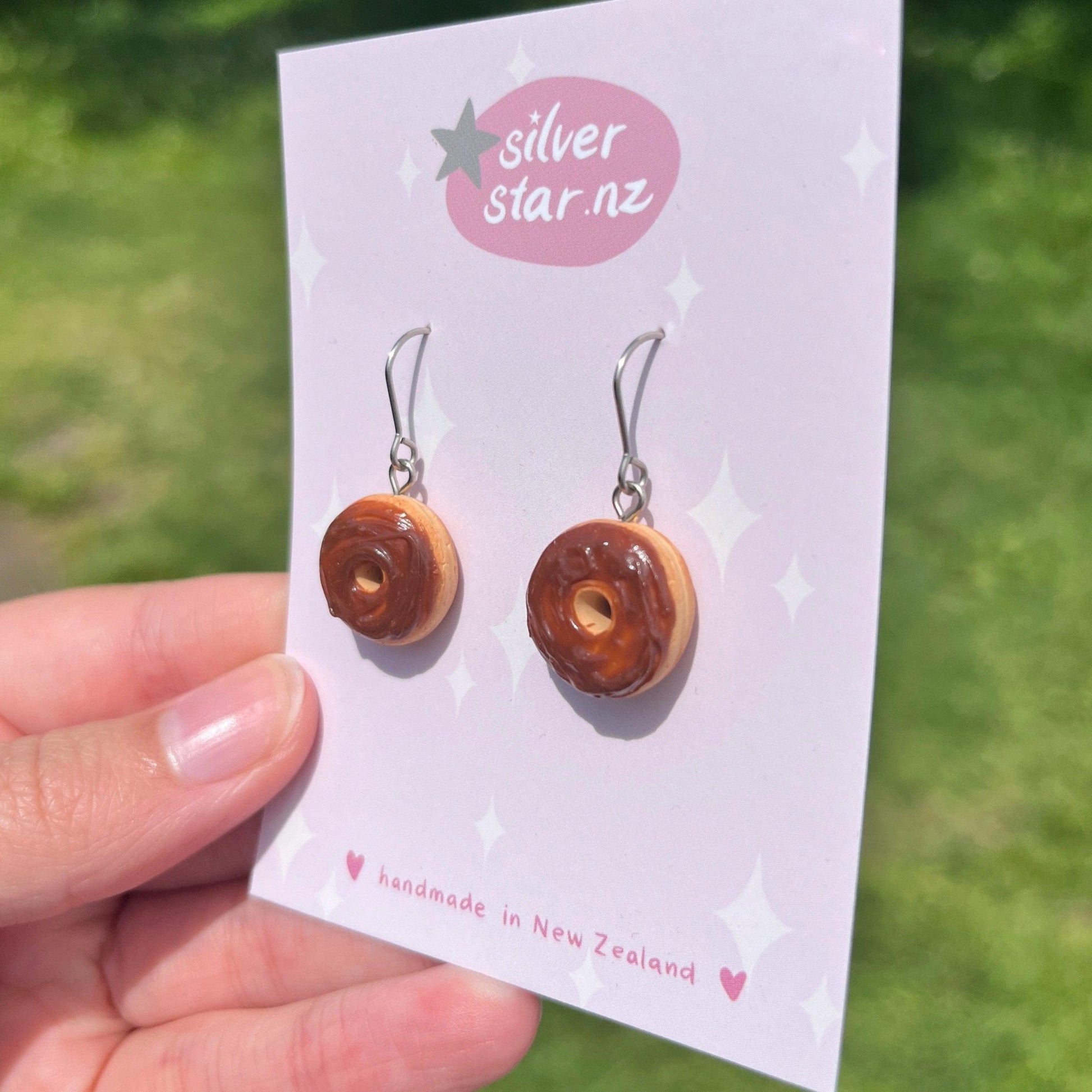 A hand displays the Choc Iced Donut Polymer Clay Earrings, each featuring delightful donut shapes with chocolate frosting, suspended from stainless steel hooks. Presented on a card labeled silverstar.nz, these handmade creations from New Zealand are set against a gently blurred green outdoor background.