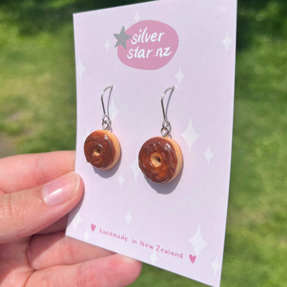 A hand displays the Choc Iced Donut Polymer Clay Earrings, each featuring delightful donut shapes with chocolate frosting, suspended from stainless steel hooks. Presented on a card labeled silverstar.nz, these handmade creations from New Zealand are set against a gently blurred green outdoor background.