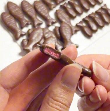 A person holding a Chocolate Fish Polymer Clay Keyring by silverstar.nz, revealing a pink filling in the center. More keyrings resembling chocolate fish are lined up on a white surface in the background, showcasing their unique design.