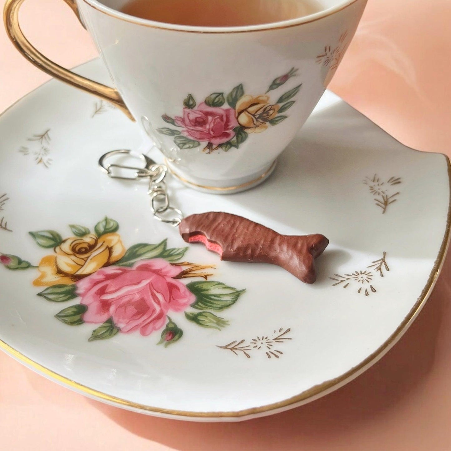 A floral teacup and saucer set, adorned with pink and yellow roses, holds a partially eaten Chocolate Fish Polymer Clay Keyring by silverstar.nz. Renowned for its unique design, the keyring is showcased on a soft pink surface bathed in sunlight.