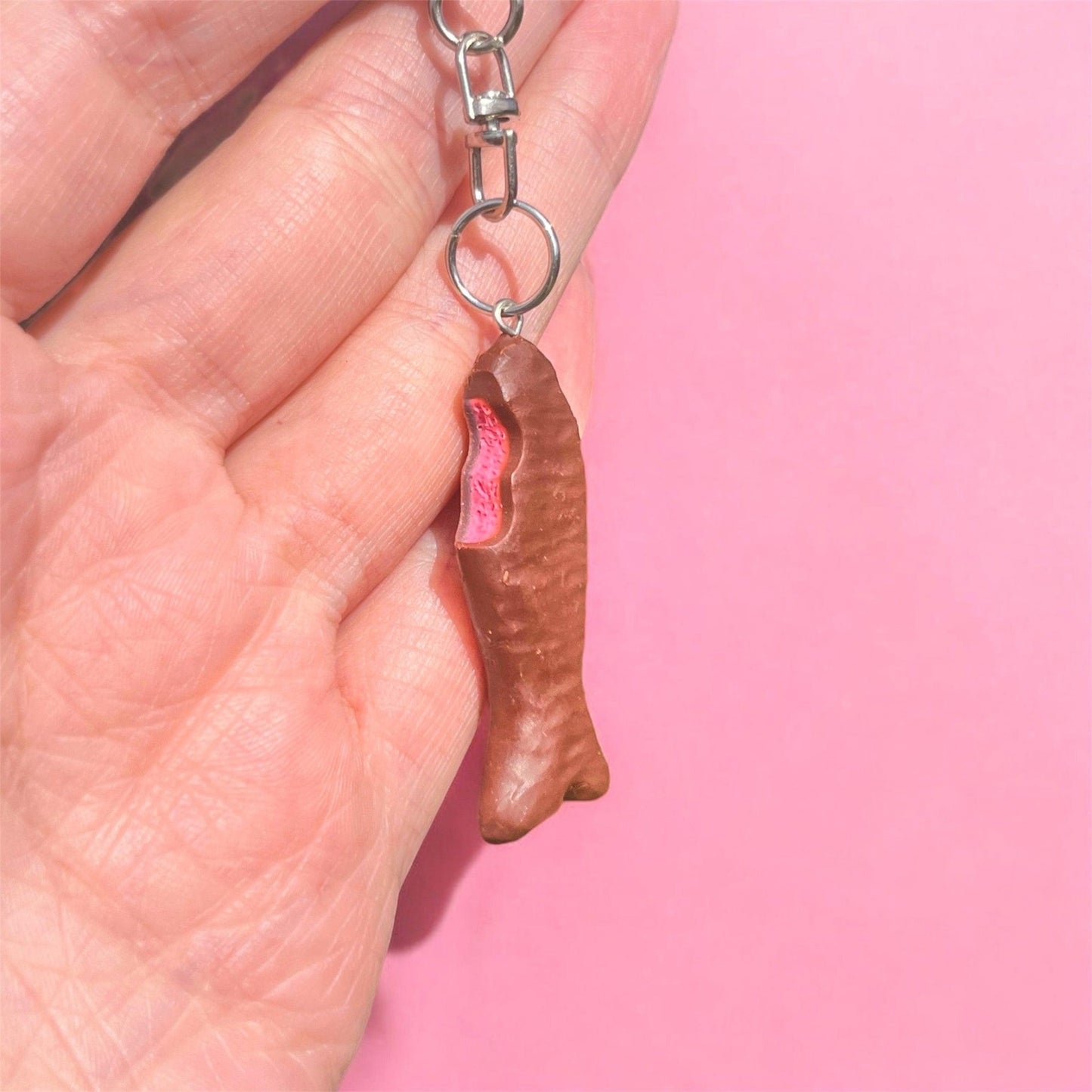 Against a pink backdrop, a hand showcases the Chocolate Fish Polymer Clay Keyring by silverstar.nz, designed to look like a bitten chocolate fish with pink filling.