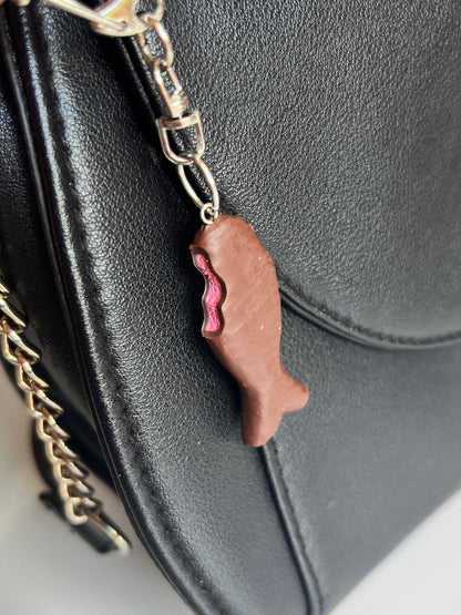 The "Chocolate Fish Polymer Clay Keyring" by silverstar.nz adorns this black leather bag with a charm that resembles a fish-shaped chocolate with pink filling. Hanging from a metal clip, the keyring beautifully complements the bag's chain strap, making it reminiscent of a unique masterpiece in keyring design.