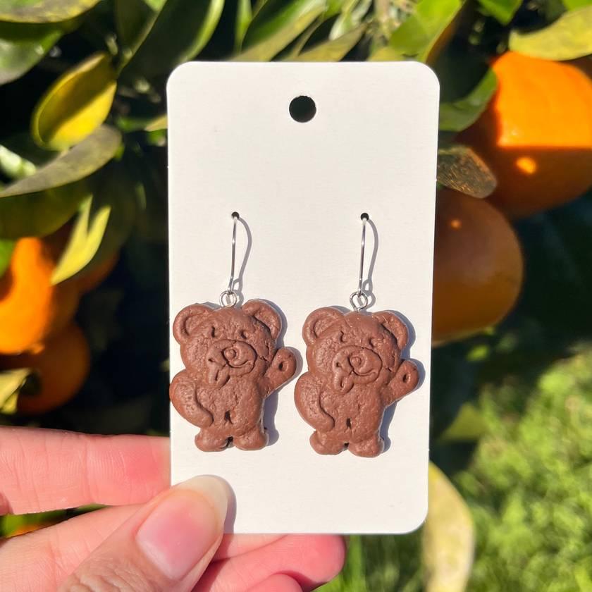 The Chocolate Tiny Teddy Polymer Clay Earrings by silverstar.nz are charming, bear-shaped accessories crafted from polymer clay and presented on a white card. They boast a cookie-like texture with a delightful brown color and are equipped with stainless steel hooks. The scene is enhanced by vibrant orange fruits and lush green leaves, evoking an inviting outdoor ambiance.