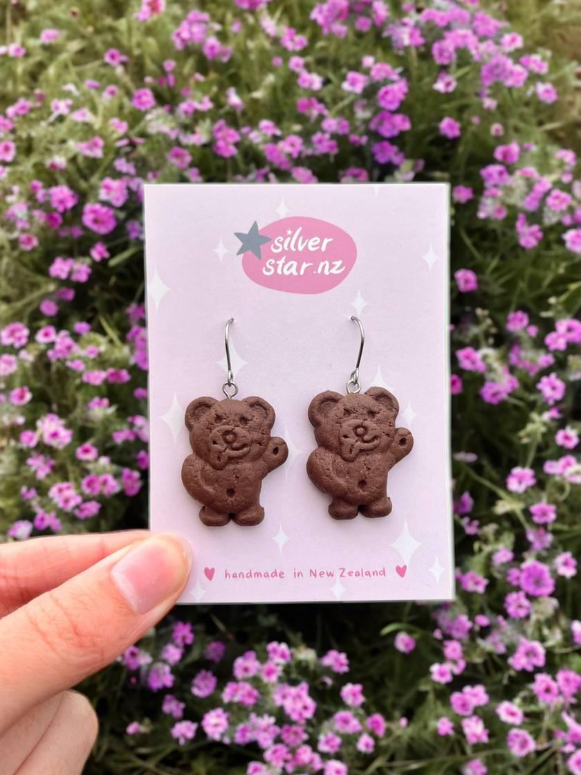 A hand displays a card featuring the Chocolate Tiny Teddy Polymer Clay Earrings against a backdrop of purple flowers. The card is labeled with "silverstar.nz" and "handmade in New Zealand," emphasizing their distinctive design and craftsmanship.