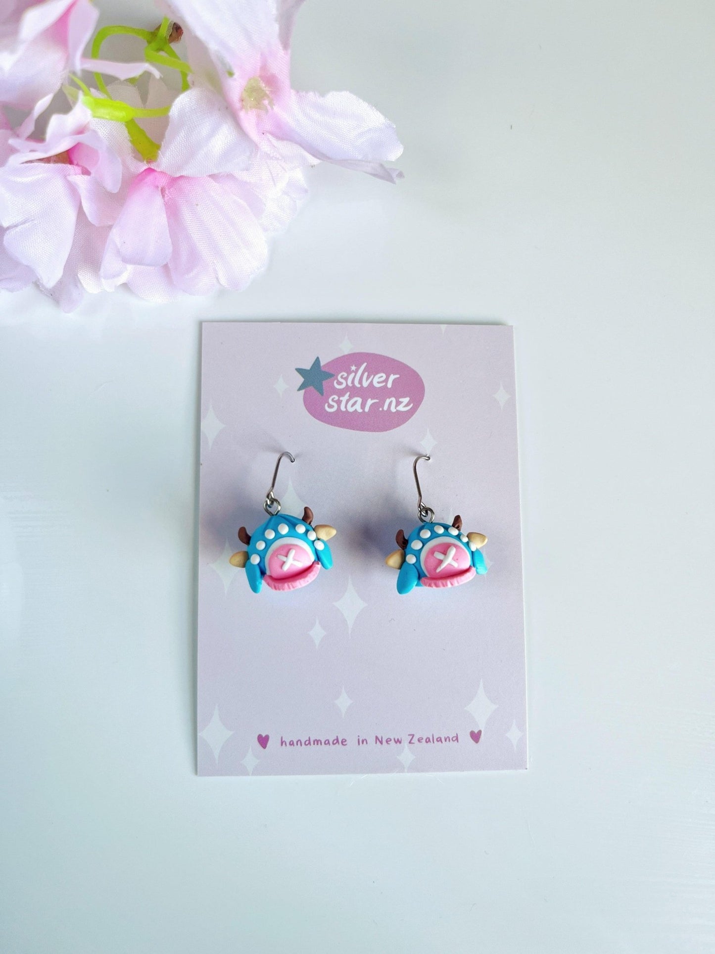 Whimsically designed in vibrant blue and pink, the Chopper Hat One Piece Polymer Clay Earrings feature unique funky designs with stainless steel hooks. Displayed on a silverstar.nz branded card stating "handmade in New Zealand," their playful charm is enhanced by peeking pink flowers in the top left corner.