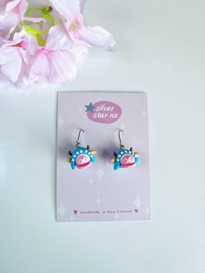 Whimsically designed in vibrant blue and pink, the Chopper Hat One Piece Polymer Clay Earrings feature unique funky designs with stainless steel hooks. Displayed on a silverstar.nz branded card stating "handmade in New Zealand," their playful charm is enhanced by peeking pink flowers in the top left corner.