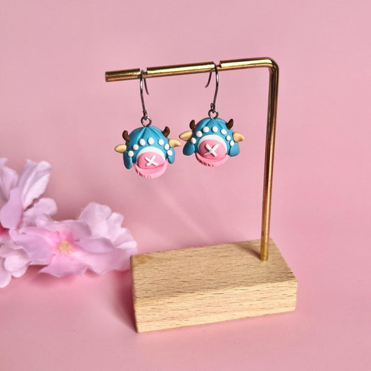 The Chopper Hat One Piece Polymer Clay Earrings by silverstar.nz showcase a unique and funky monster-inspired design, handcrafted with pink and blue shades, playful horns, and spots. They are displayed on a gold stand with stainless steel hooks and adorned by a pink flower resting beside the wooden base against a pastel pink backdrop.