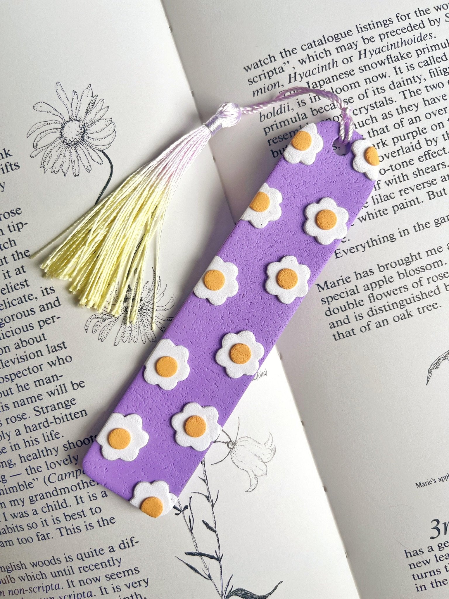 A Daisy Polymer Clay Bookmark by silverstar.nz, colored in purple and embellished with white and yellow daisy motifs, elegantly rests on an open book showcasing botanical illustrations. This handmade bookmark, ideal as a gift for a loved one, features a light yellow tassel for added charm.