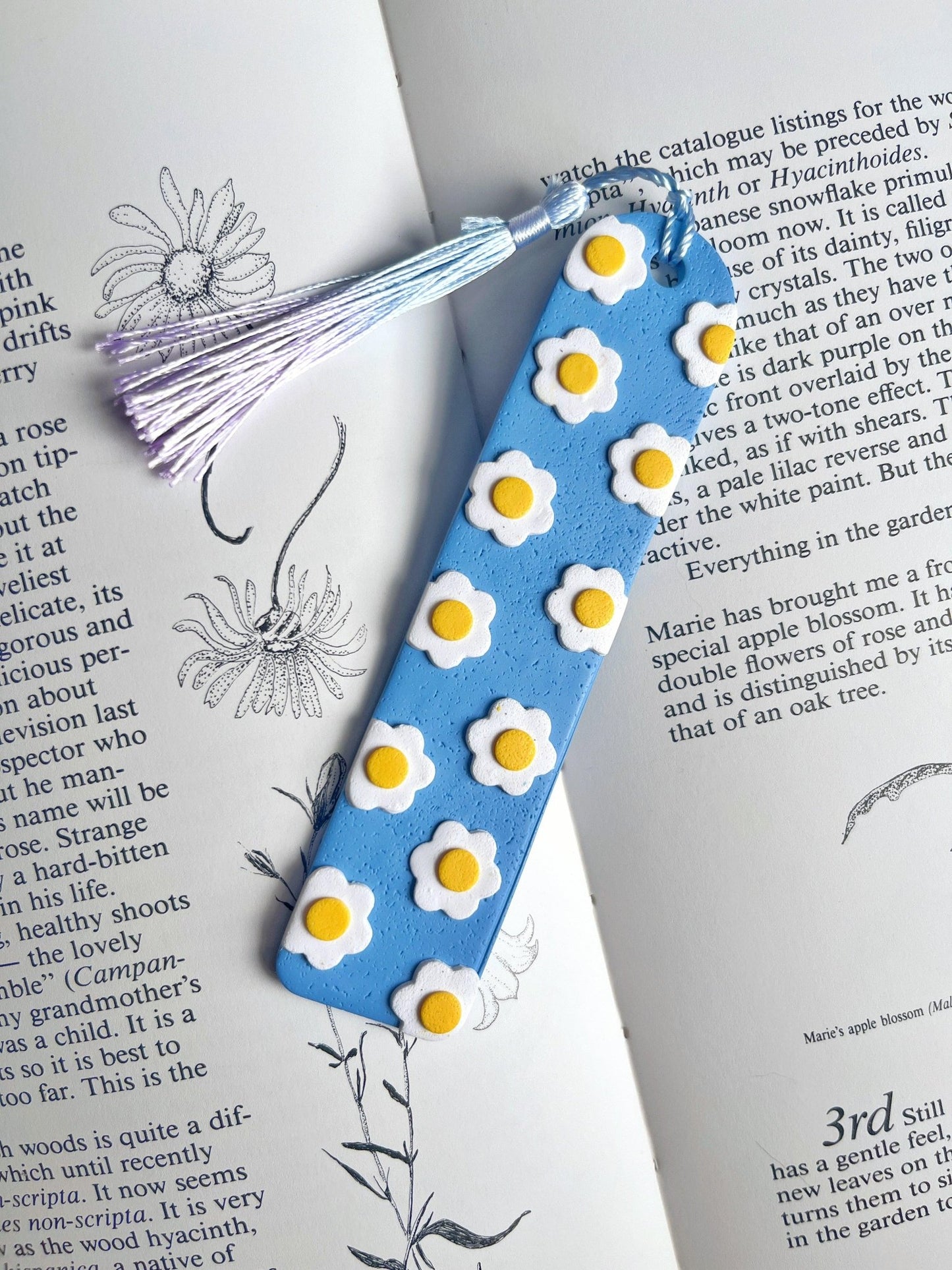 A handmade Daisy Polymer Clay Bookmark from silverstar.nz, featuring blue polymer clay adorned with white and yellow daisy-like flowers, lies on an open book filled with botanical illustrations and text. Attached is a lavender tassel, making it the perfect gift for a loved one.