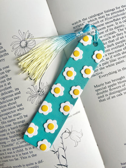 A Daisy Polymer Clay Bookmark from silverstar.nz, adorned with white and yellow daisy designs on a turquoise background, lies on an open book, making it a perfect handmade gift for a loved one. The bookmark is completed with a tassel in shades of white, blue, and yellow. The book's pages are enriched with text and floral illustrations.