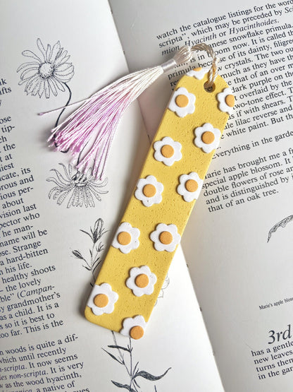 A Daisy Polymer Clay Bookmark from silverstar.nz, featuring handmade yellow designs with white and orange flowers, is placed on an open book of botanical illustrations and text. This thoughtful gift for a loved one is completed with a white and pink tassel.