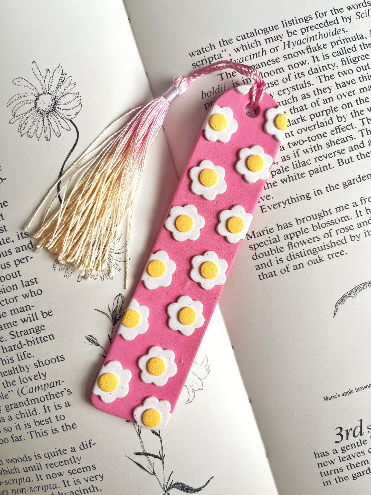 A pink handmade Daisy Polymer Clay Bookmark from silverstar.nz, adorned with small daisies and fried egg designs, rests on an open book. Its white and pink tassel adds charm to the scene. The book's pages display botanical illustrations and text, making it a perfect gift for a loved one who cherishes nature.