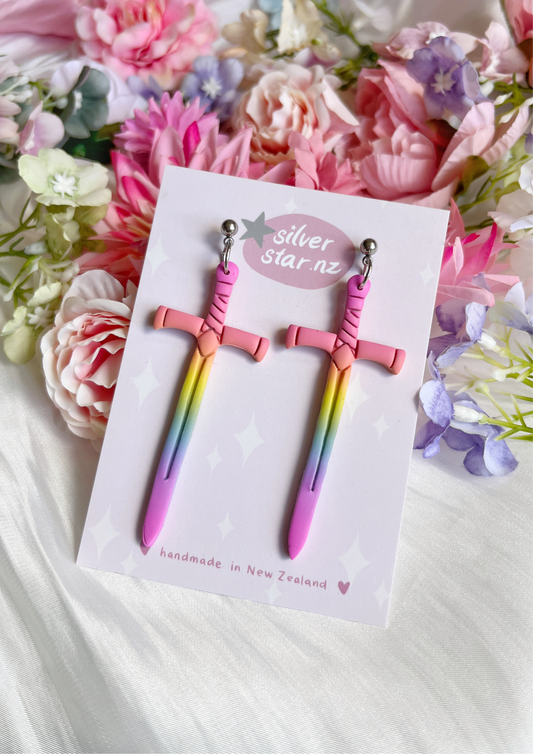 The Sword Polymer Clay Earrings, designed by silverstar.nz, are colorful sword-shaped handmade earrings with a pink, yellow, and purple gradient. Theyre displayed on a silverstar.nz card against a pastel floral background that highlights their unique design.