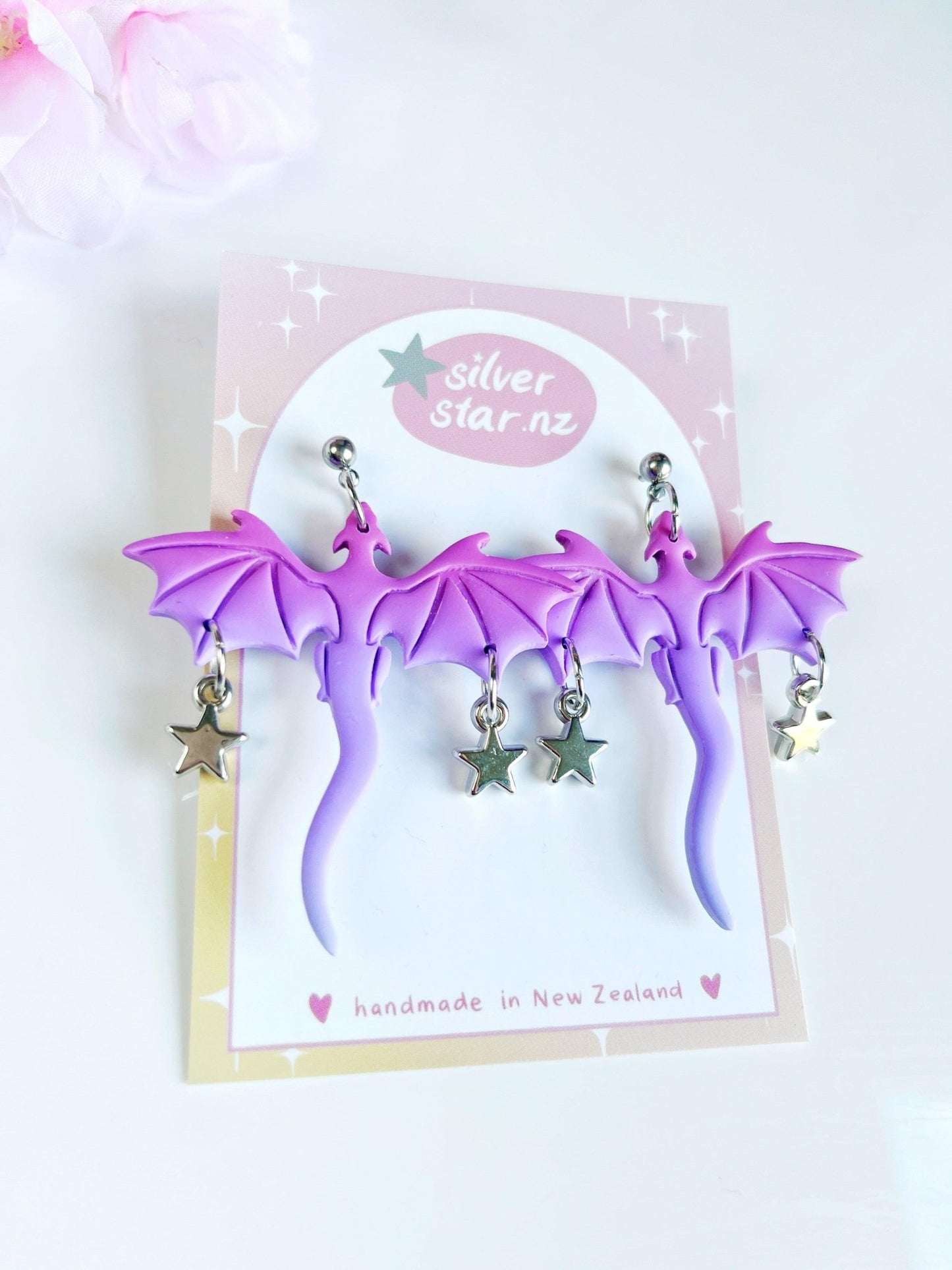 A pair of unique Purple Dragon Polymer Clay Earrings with dangling silver stars is displayed on a card labeled silverstar.nz and handmade in New Zealand. A pink flower is partially visible in the background, accentuating these exquisite handmade earrings.