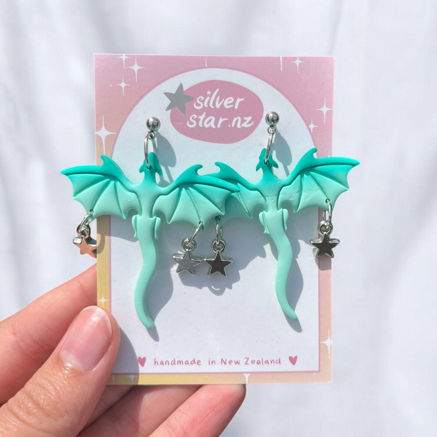 A hand holds a card displaying the (Imperfect) Mint Dragon Polymer Clay Earrings, adorned with wings and intricate silver star charms. The pink card, featuring a rainbow gradient border and the brand name "silverstar.nz," emphasizes that these exquisite earrings are handmade in New Zealand.