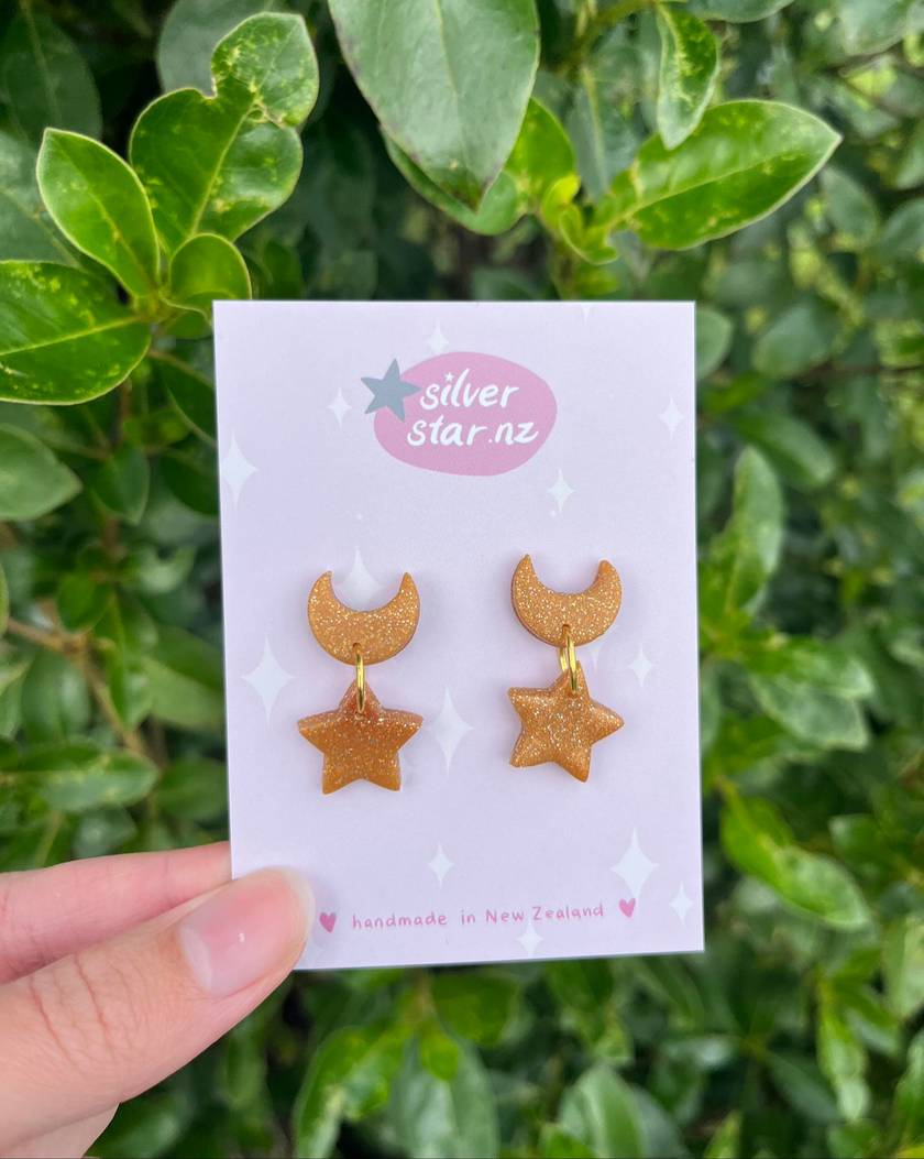 A hand displays a card featuring Star + Moon Polymer Clay Earrings from silverstar.nz, adorned with gold glittery moons and stars. The backdrop of green leaves complements the cards message: handmade in New Zealand with heart details, celebrating the charm of handcrafted earrings.