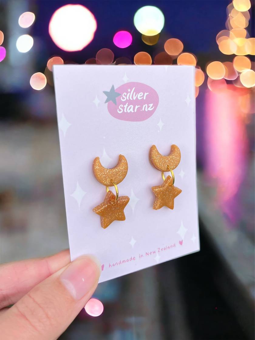 A hand presents Star + Moon Polymer Clay Earrings by silverstar.nz on a branded card. The design, featuring glittery gold crescent moons and stars, stands out against a blurred nightscape with colorful bokeh lights reflecting on water, highlighting the earrings whimsical charm.