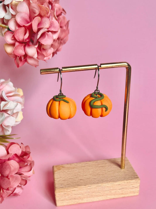 The Pumpkin Polymer Clay Earrings, featuring a distinctive mini pumpkin design, are displayed on a gold stand with a wooden base. These handmade earrings are set against a pink backdrop embellished with clusters of pink flowers on either side.